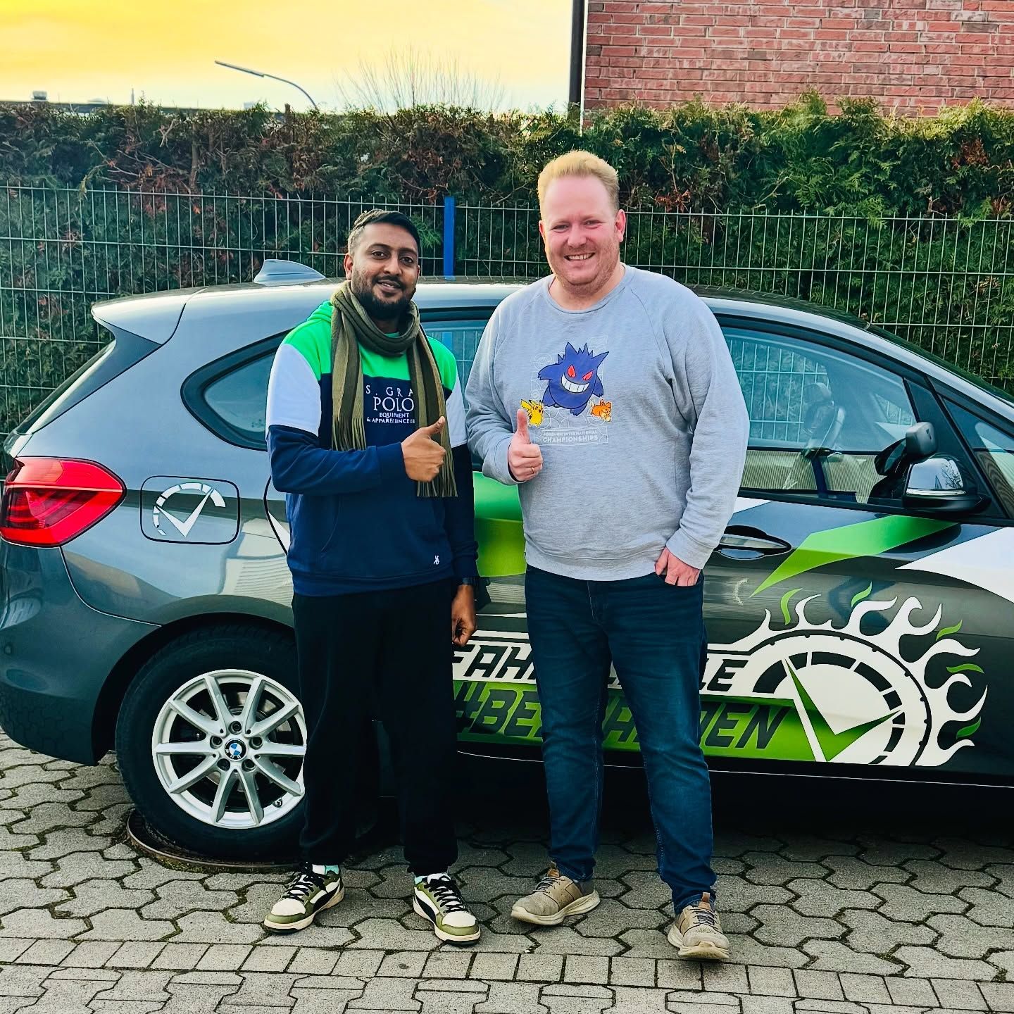 Ein Fahrschüler und Fahrlehrer Lukas zeigen vor dem Fahrschulwagen den Daumen nach oben