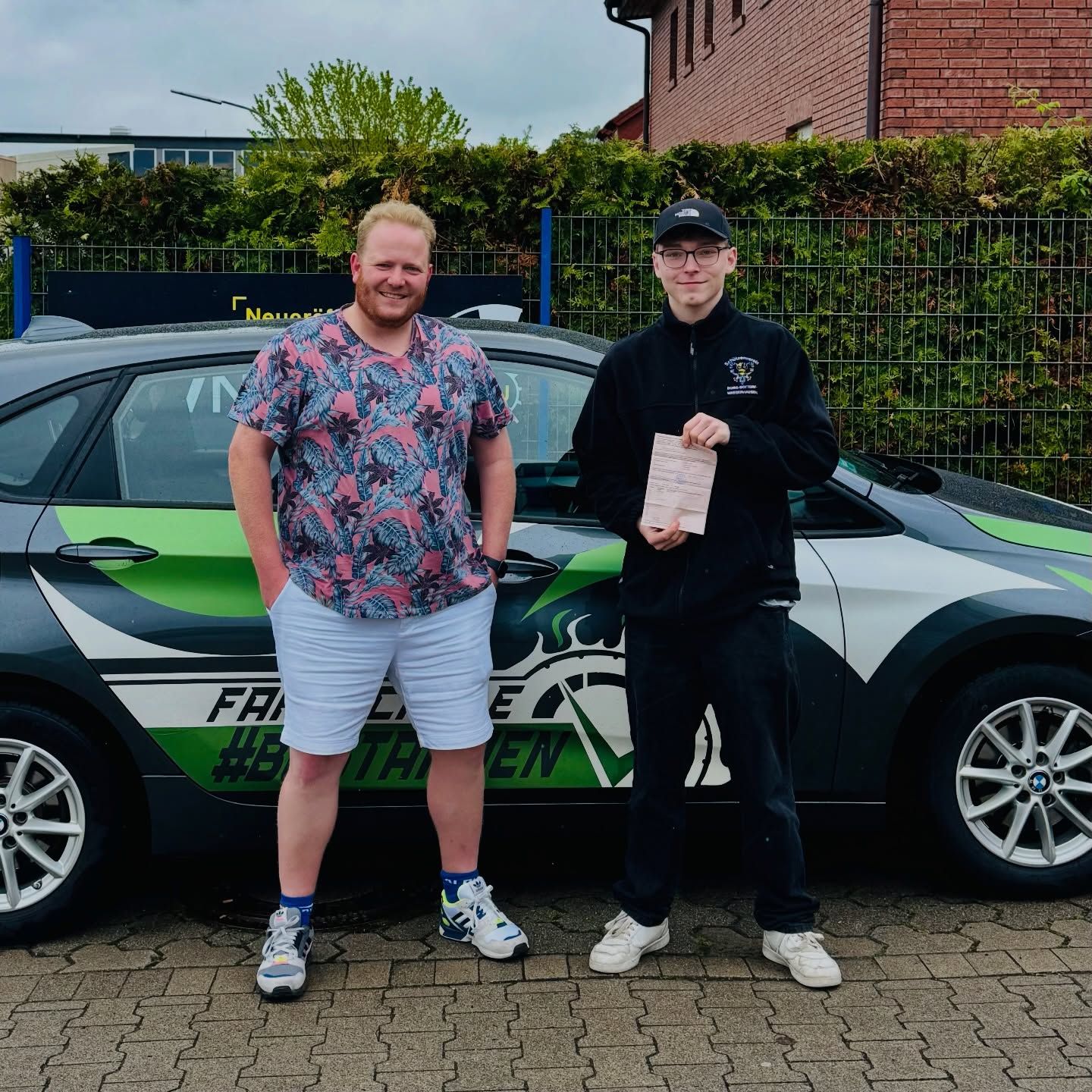 Fahrlehrer Lukas und ein Fahrschüler mit Prüfungsbescheinigung vor dem Fahrschulwagen