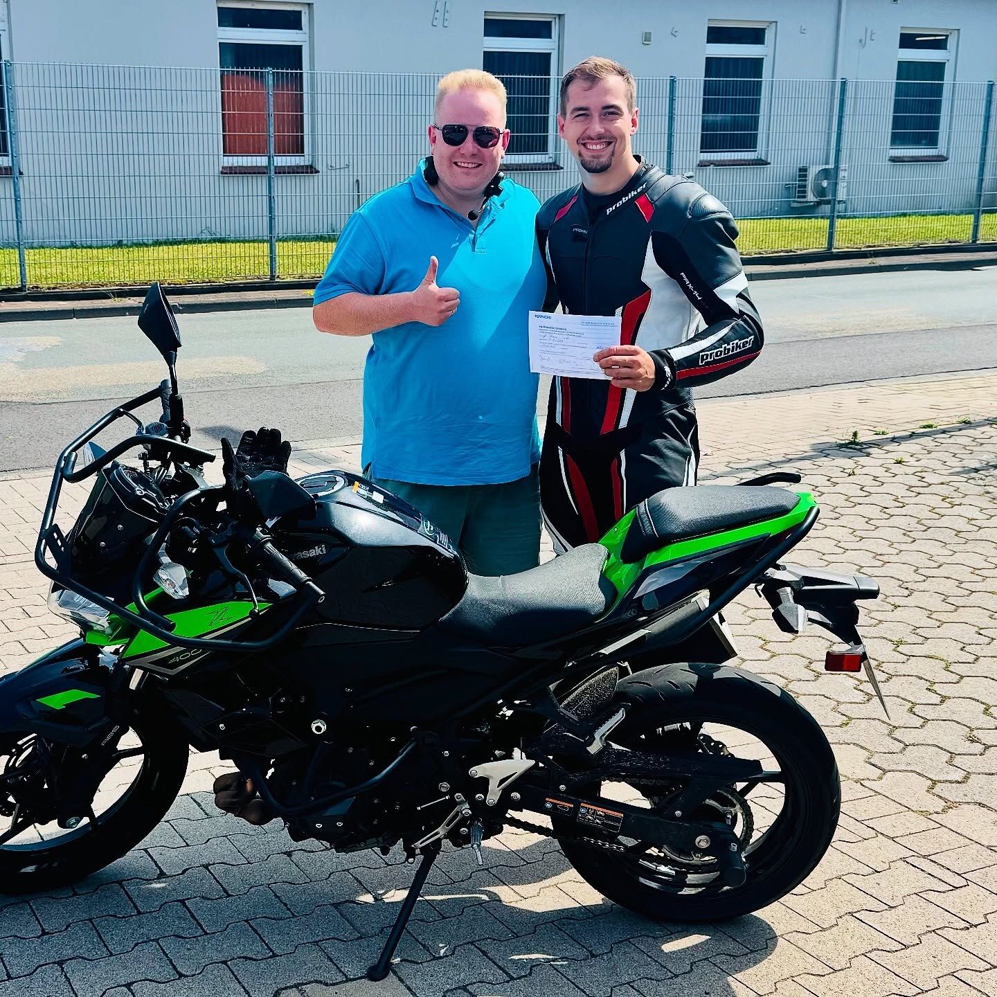 Fahrlehrer Lukas und ein Fahrschüler im Motorradanzug mit dem Prüfungszeugnis neben dem Motorrad